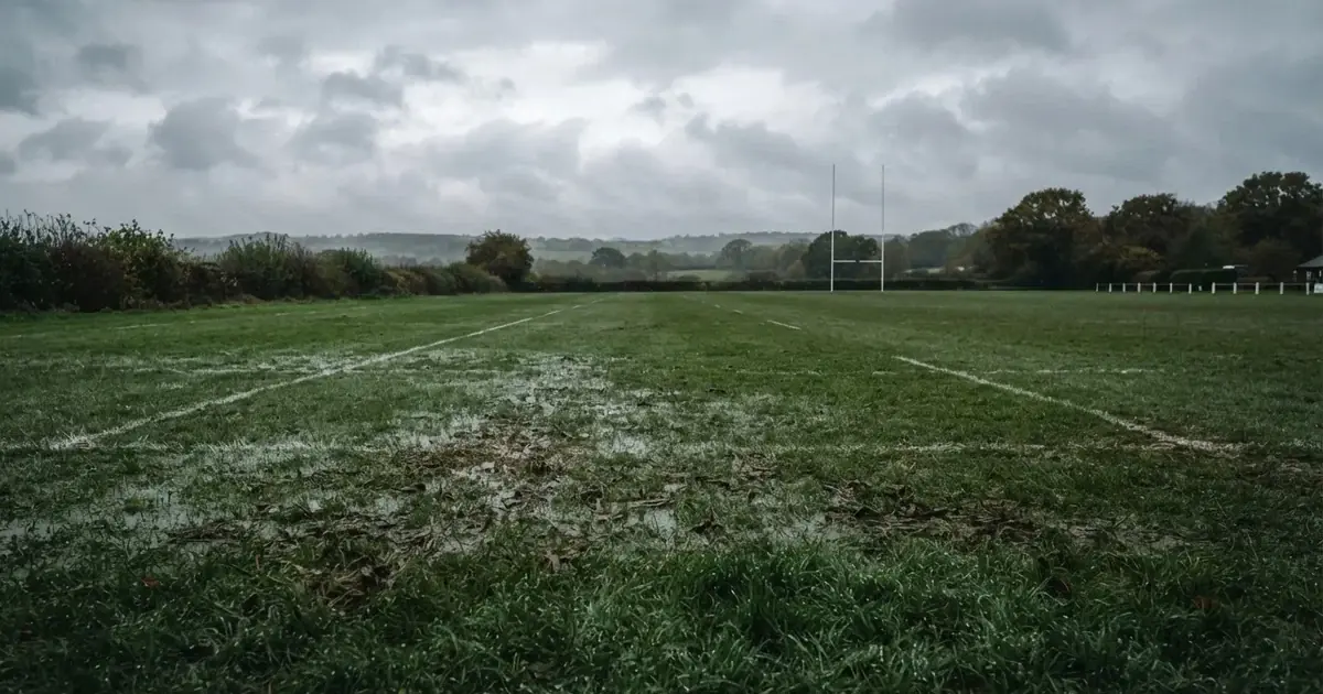 Boisko do rugby ze słupami bramki w oddali pod pochmurnym niebem, mokra trawa i ślady butów.