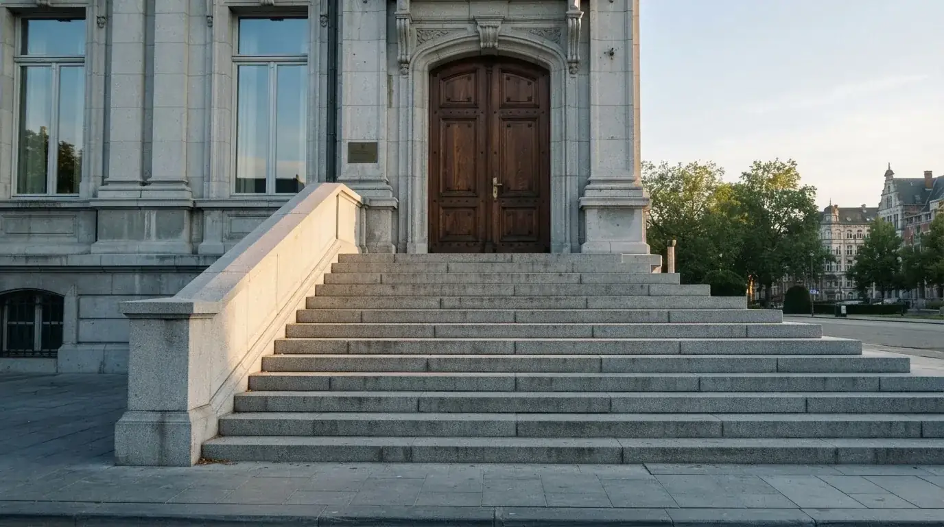 Granitowe schody prowadzące do wysokich drewnianych drzwi budynku w porannym świetle, pusta balustrada.