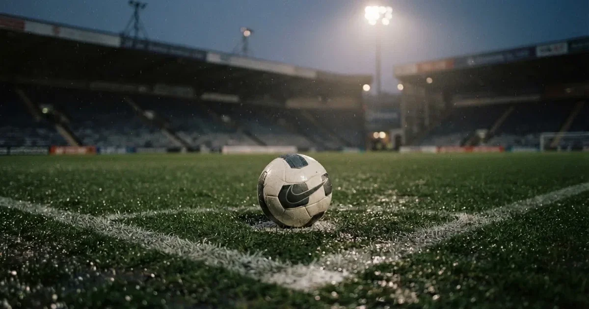 Futbolowa piłka stoi na punkcie karnym w pustym stadionie podczas mżawki, w tle rozmyte trybuny i jasne światło masztów.