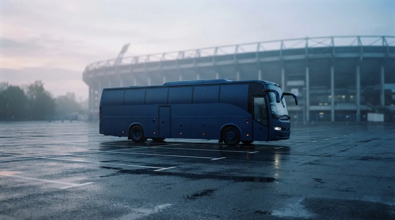 Autokar stoi na parkingu przed stadionem o świcie, rozmyte kontury trybun w tle, pusty plac i mokry asfalt.